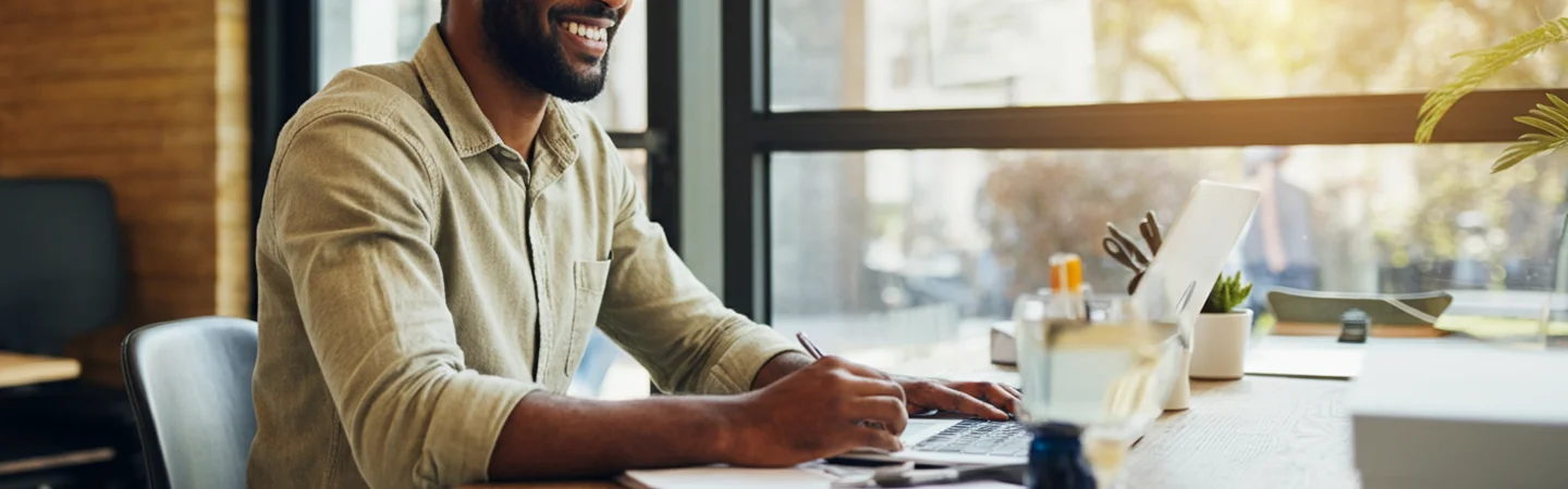 A man sits at a desk, focused on his laptop, with papers and a coffee cup nearby.
