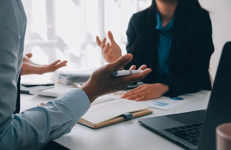 Two professionals engaged in a discussion at a desk with a laptop, notebook, and documents.