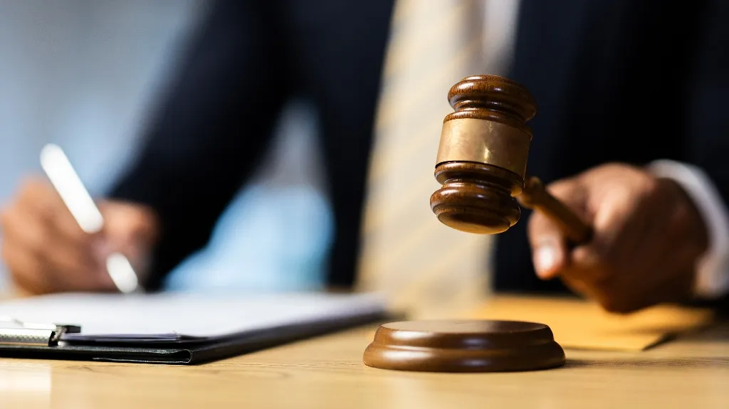 Close-up of a judge’s hand holding a wooden gavel above a sound block with blurred person writing on clipboard in background.