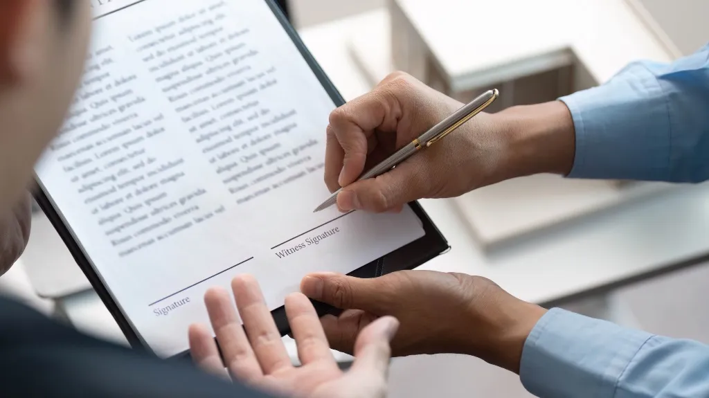 Person in blue shirt signing a formal document labeled with signature and witness signature lines.