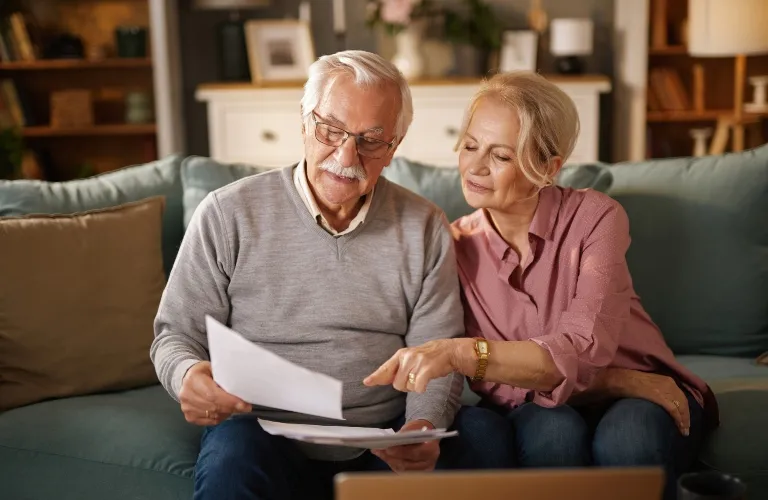 Elderly couple sitting on sofa reviewing documents together in a warmly lit living room.