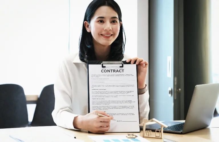 Professional woman in white blouse holding and presenting a contract document at office desk.